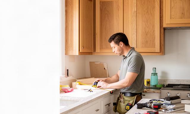 Renovation professional measuring a bright kitchen beside oak cabinets and a stone countertop